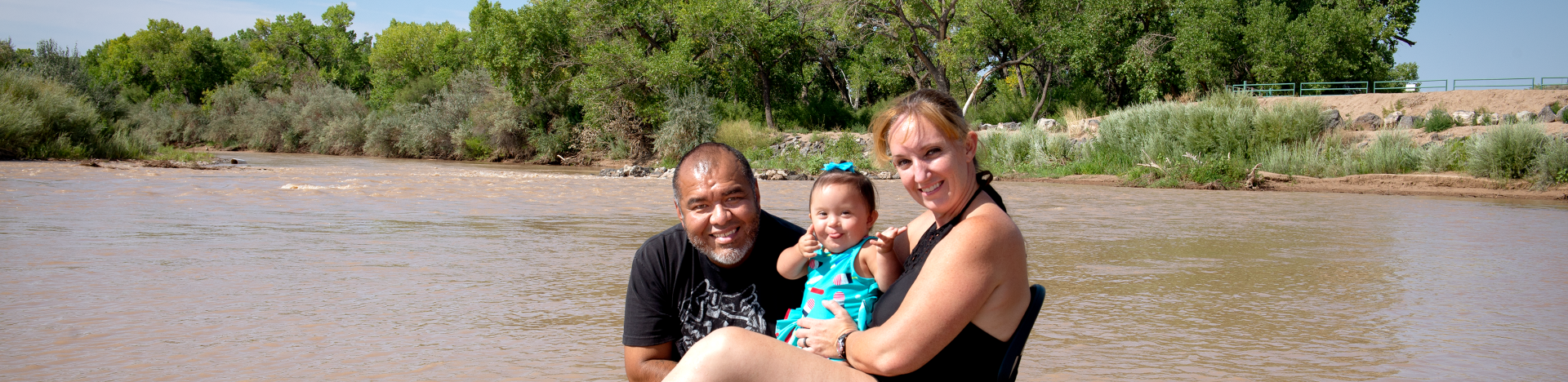 Parents with their toddler by smiling and swimming by the river.