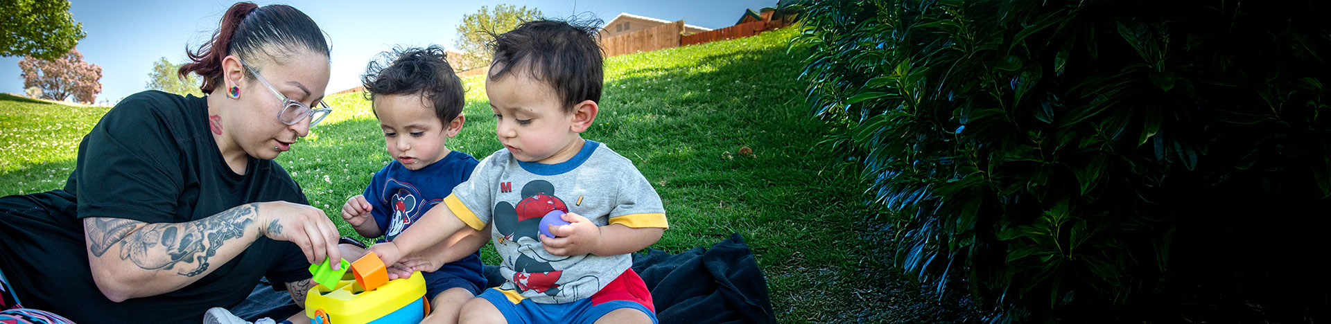 a mother sitting in the grass with her two children playing with a puzzle toy