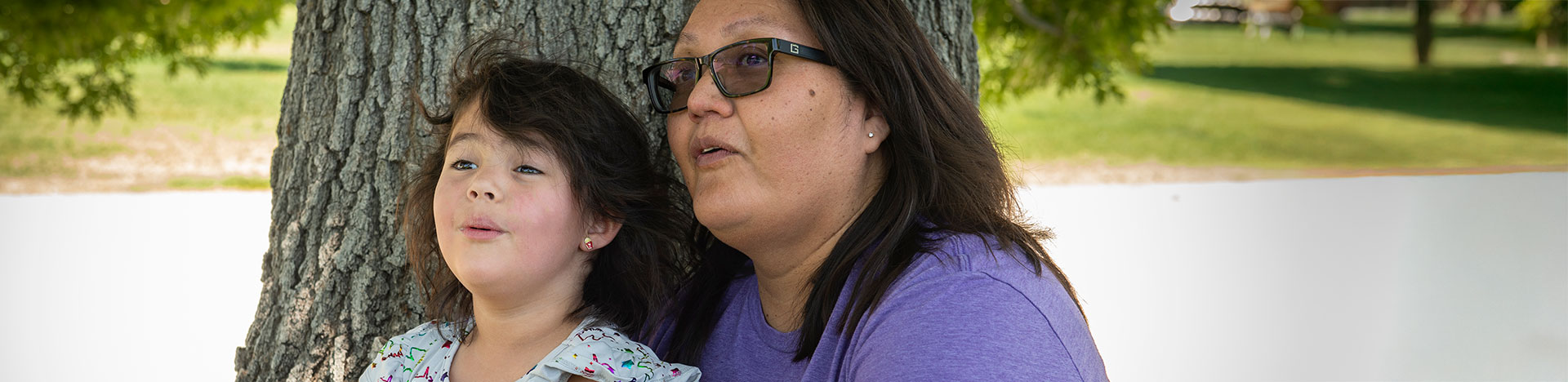 A young girl sitting with her mother under a tree and they're both looking at something in the distance