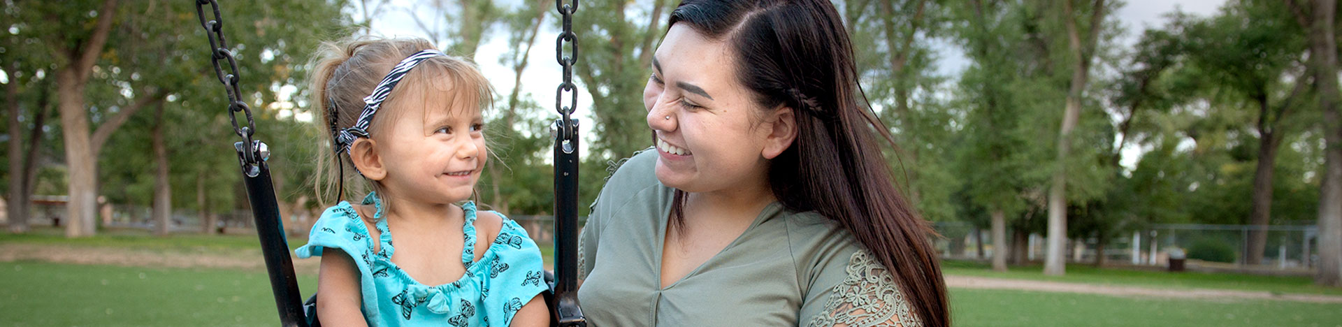 A mother smiling at her daughter who is swinging at a park.